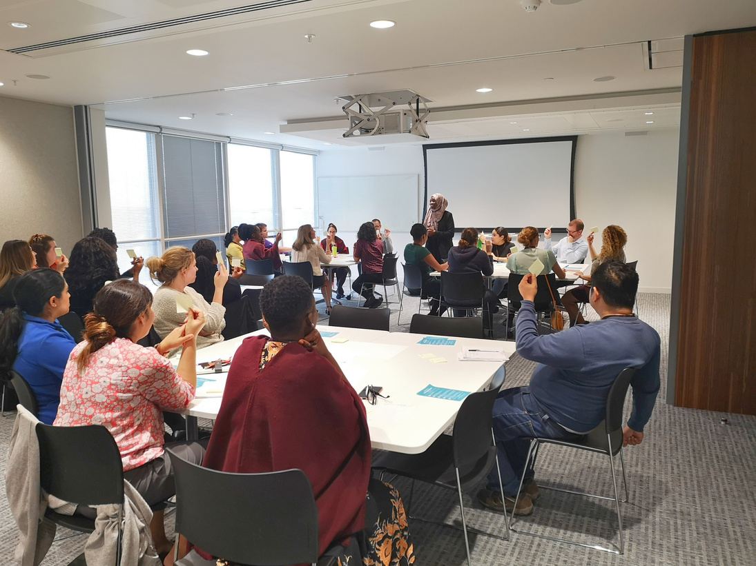 A full workshop room of leaders seated at round tables, holding up cards